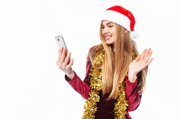 Beautiful girl in the hat of Santa Claus and dress, with a smartphone in his hands and looking at him, sends greetings. On a white background - Photo, image