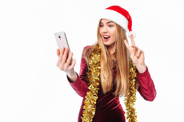 Beautiful girl in the hat of Santa Claus and dress, with a smartphone in his hands and looking at him, sends greetings. On a white background - Photo, Image