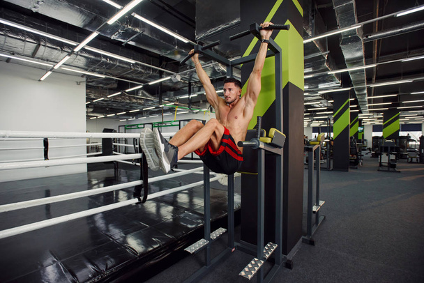 Strong healthy guy doing abdominal exercises on the crossbar during training in a modern gym with black and green interior. - Фото, изображение