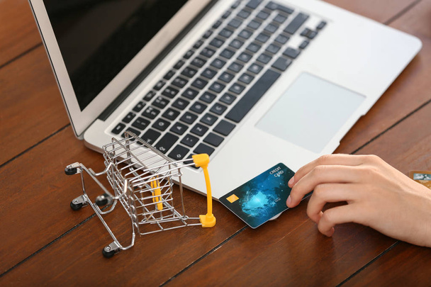 Frau mit Kreditkarte, kleiner Einkaufswagen mit Laptop auf Holztisch, Nahaufnahme. Online-Einkauf - Foto, Bild