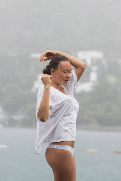 Çekici genç bir kadın bluz ve külot giyerken kendini kucaklıyor ve rüzgarlı bir günde suyun yanında dururken gözlerini kapalı tutuyor. - Fotoğraf, Görsel