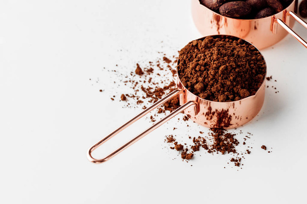Oro rosa que mide tazas de granos de cacao, nabos de cacao y cacao en polvo sobre un fondo blanco, concepto de alimentos saludables - Foto, imagen