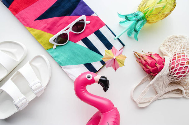 Flat lay of summer vibes concept with colorful travel fashion items, sunglasses, scarf, pink dragon fruit, flamingo inflatable drink holder, pineapple straw tumbler bottle, net bag and sandal on a white background. - Photo, Image