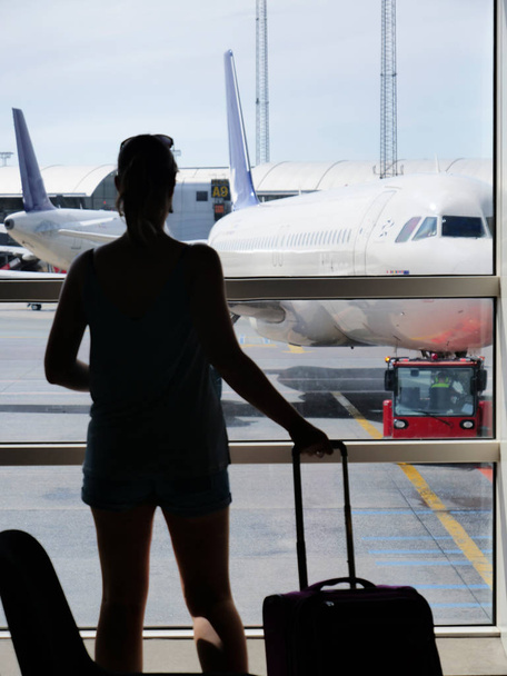 SILHOUETTE: Woman grabs her luggage and looks through the window at a large airplane parked in the sunny runway. Unrecognizable female traveler holding her travel bags while observing the airport. - Photo, Image