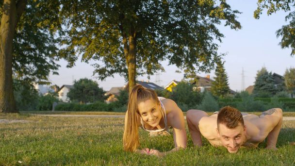 Portret: Shirtless man met tattoo doet push-ups in de met gras begroeide park terwijl lachende vrouw houdt een plank. Energetische jong stel samen op zonnige aard uitoefenen. Gelukkig man en vrouw opleiding buiten - Foto, afbeelding