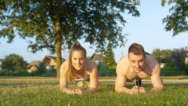 PORTRAIT : Couple sportif souriant tenant un exercice de planche dans le parc verdoyant ensoleillé. Jeune petit ami caucasien et sa petite amie exerçant leur noyau dans la nature tranquille lors d'une belle soirée d'été
. - Photo, image