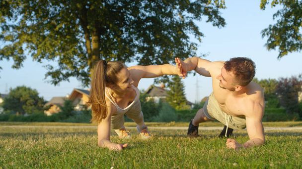 CLOSE UP: Cheerful sporty couple working out in the park hold a plank and high five while turning to the side. Young athletic Caucasian boyfriend and girlfriend enjoying summer by training outside. - Photo, Image