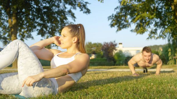 SLOW MOTION, CLOSE UP: Fit young Caucasian couple working out together in the sunny nature. Fit woman doing crunches while shirtless boyfriend does press ups in background. Athletes training outside. - Фото, зображення