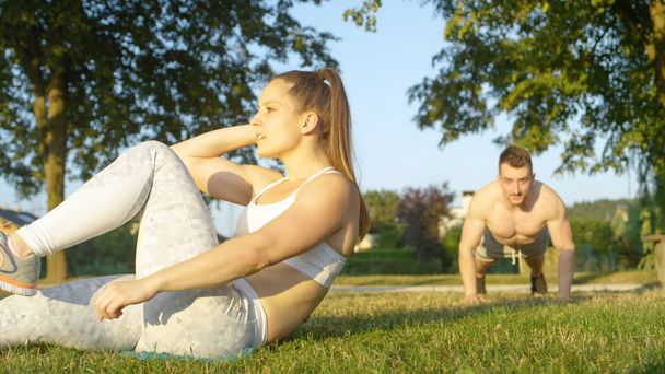 SLOW MOTION, PORTRAIT: Smiling couple having fun working out together in the sunny park. Cheerful young woman does sit ups while shirtless boyfriend does push ups in green nature during the summer. - Φωτογραφία, εικόνα