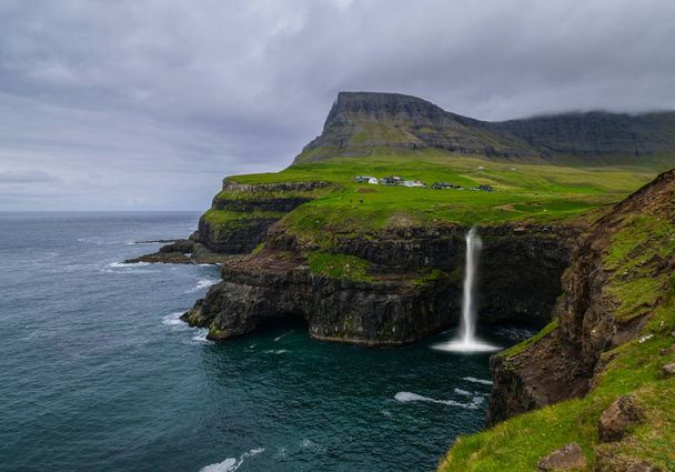 Antenne: malerisches Dorf unter grasbewachsenen Bergen und den spektakulären Klippen an einem bewölkten Tag auf den atemberaubenden Färöer-Inseln. schöner Blick auf einen spektakulären Wasserfall in skandinavischer Landschaft. - Foto, Bild