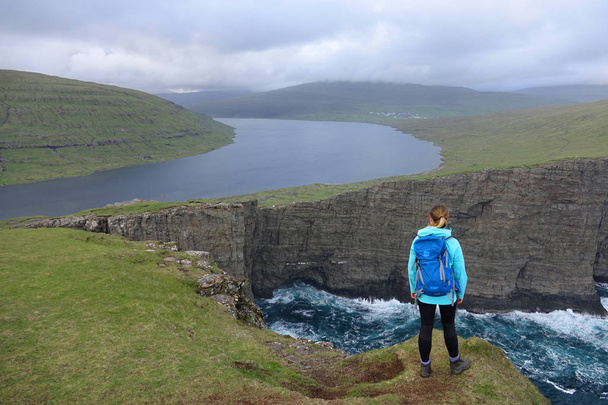 Antenne: Eine junge Reisende wandert bis an den Rand einer Klippe, um die atemberaubende grüne Landschaft der stimmungsvollen Färöer-Inseln zu sehen. Wanderin in Skandinavien beobachtet stillen See und Wellen, die auf Felsen krachen - Foto, Bild