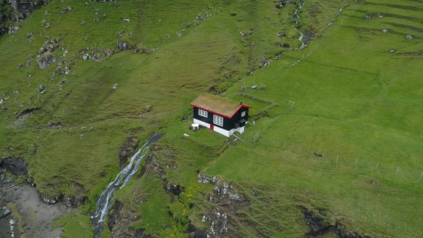 Εναέρια: Πετούν πάνω από ένα απομακρυσμένο σπίτι στην τραχιά σκανδιναβική ύπαιθρο περιβάλλεται από απότομους χλοώδεις λόφους. Καταπληκτική θέα του βουνού ρεύμα που ρέει μέσα σε μια λίμνη κοντά στο μοναχικό μαύρο σπιτάκι. - Φωτογραφία, εικόνα