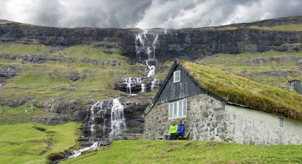 Young man and woman sit on a wooden bench by the stone house near the breathtaking waterfalls right before a storm. Young tourist couple sitting by the traditional grassy house in Faroe Islands. - Photo, Image