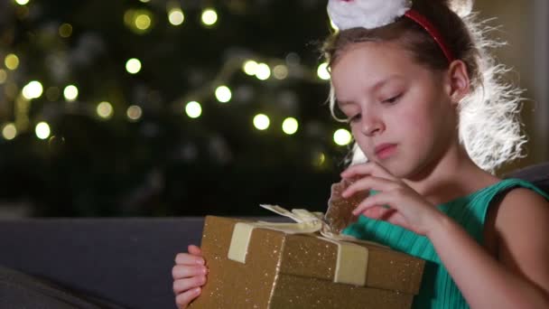 Happy child opening Christmas gift box. Funny girl dressed in Santa Claus hat in bedroom. Portrait of smiling kid at home. Xmas holiday concept - Imágenes, Vídeo