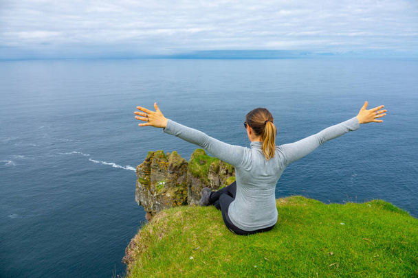 Eine unkenntliche Frau sitzt mit ausgestreckten Armen auf dem grasbewachsenen Rand einer Klippe und blickt in Richtung offene See. spektakuläre Aussicht auf den endlosen Ozean von einer hohen Klippe auf den spektakulären Färöer-Inseln - Foto, Bild