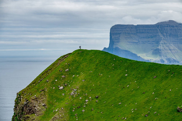 Caminante masculino irreconocible se encuentra en la colina cubierta de hierba en la distancia y toma fotos de fiordos impresionantes en las Islas Feroe. Hermosa foto de un hombre pequeño en el magnífico campo escandinavo
. - Foto, imagen