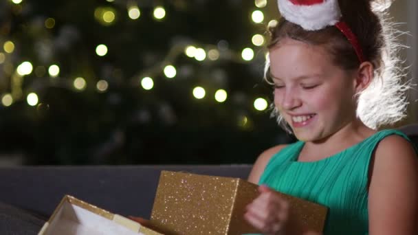 Sweet girl rejoices at a Christmas present. Schoolgirl is sitting on the sofa opposite the New Year tree hugging a box with a gift. The girl wears a decorative hoop with a Santa hat - Πλάνα, βίντεο