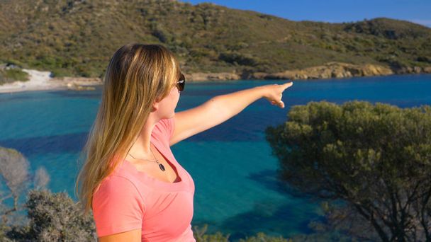 Κοντινό πλάνο: Άγνωστη νεαρή γυναίκα σημείο προς τη γραφική παραλία από άποψη πάνω από τον γαλάζιο κόλπο σε απομακρυσμένο νησί της Ιταλίας. Αγνώριστος θηλυκό τουριστών που δείχνει η γραφική ακτή της Σαρδηνίας. - Φωτογραφία, εικόνα