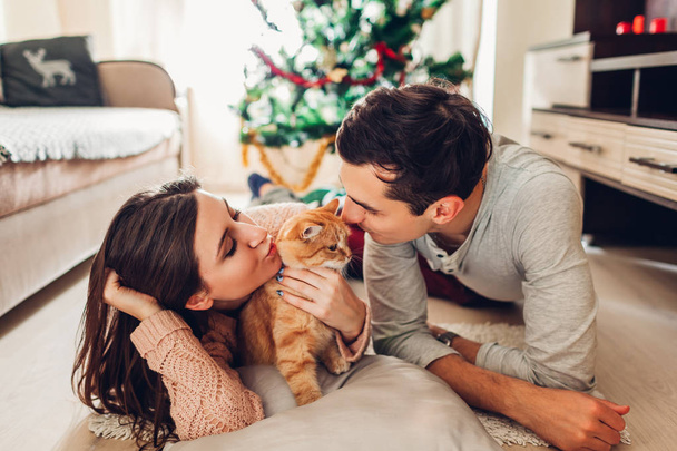 Couple in love lying by Christmas tree and playing with cat at home. Happy man and woman kissing pet - Foto, imagen