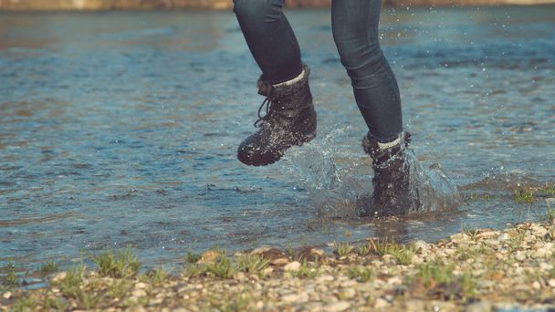 LOW ANGLE: Tunnistamaton leikkisä nainen pörröinen saappaat nauttia syksyllä juoksemalla ulkona pitkin matalaa virtaa. Energinen tyttö hauskaa roiskeita järven vesi hohtavan lämpimässä auringossa
. - Valokuva, kuva