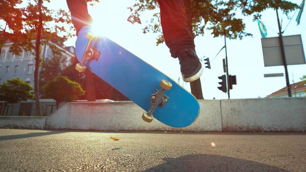 クローズアップ、レンズフレア:認識できない男性アスリートは、公園で晴れた日に彼の青いスケートボードでfakieトリックを行います。若いスケーターは、舗装を転がり、彼のデッキでフリップを行います. - 写真・画像