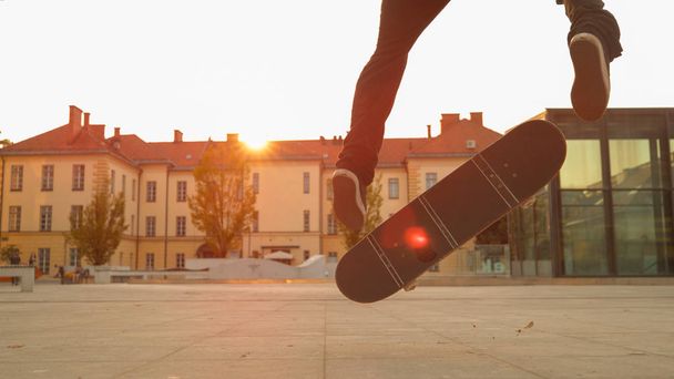 LENS FLARE, CLOSE UP, LOW ANGLE: Jovem patinador pousa um truque legal enquanto no skateboaring no parque urbano ao pôr do sol idílico. Homem skatista faz um fakie durante uma sessão à noite em uma praça da cidade
. - Foto, Imagem