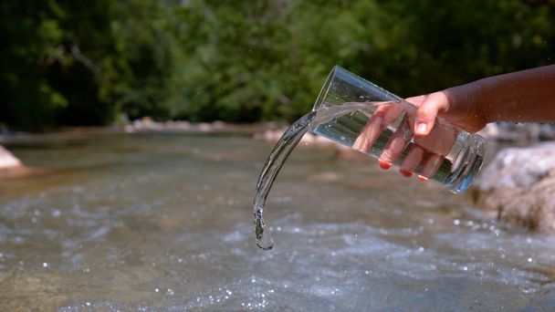クローズ アップ: 認識できない若い人は静かな山川に大きな透明ガラスから透明な水を注ぐ。ガラス液体の流出ストリームにハイカーの少女の映画のようなショット. - 写真・画像