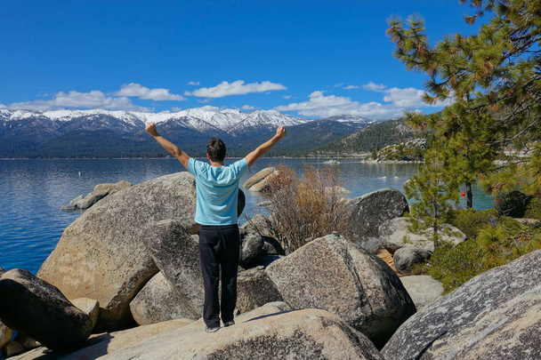 Unrecognizable Caucasian man celebrates reaching the breathtaking Lake Tahoe on a spectacular sunny day. Picturesque wilderness surrounds unknown male hiker standing on a boulder with his arms raised - Photo, Image