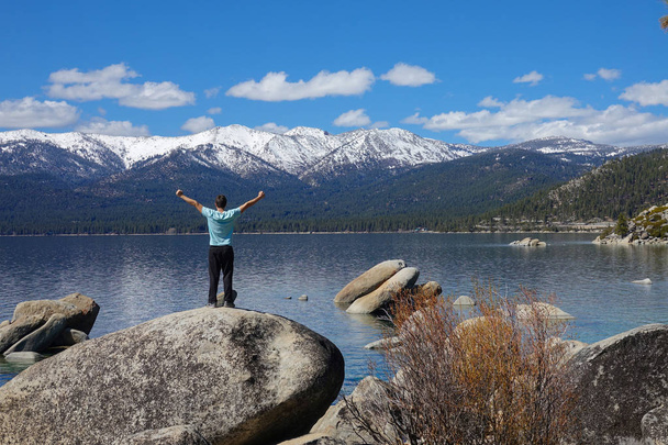 Jeune homme inconnu randonnée dans la nature californienne célèbre tout en se tenant sur un rocher dans le lac Tahoe pittoresque. Touriste masculin aux bras tendus observe les montagnes enneigées et le grand lac
. - Photo, image