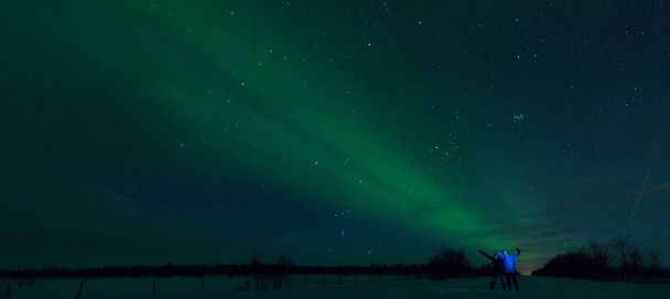 Jonge Kaukasische toeristische paar outstretches wapens en viert de spectaculaire aurora borealis te zien. Gelukkig vriend en vriendin plezier observeren het adembenemende kleurrijke Noorderlicht. - Foto, afbeelding