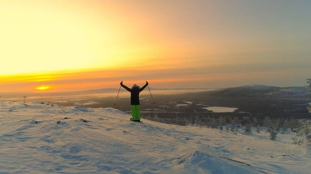 Antenne: Eine unkenntliche Skifahrerin blickt bei Sonnenuntergang über das spektakuläre Lappland. Eine junge Frau feiert an einem sonnigen Winterabend das Erreichen des Gipfels auf ihren Skiern. malerische winterliche Natur in Finnland - Foto, Bild