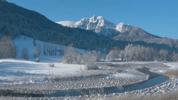 Spectacular mountains in Slovenia tower over the snowy forest and small stream running through the idyllic sunny countryside. Breathtaking view of the snowy spruce forest and beautiful rocky mountains - Photo, Image