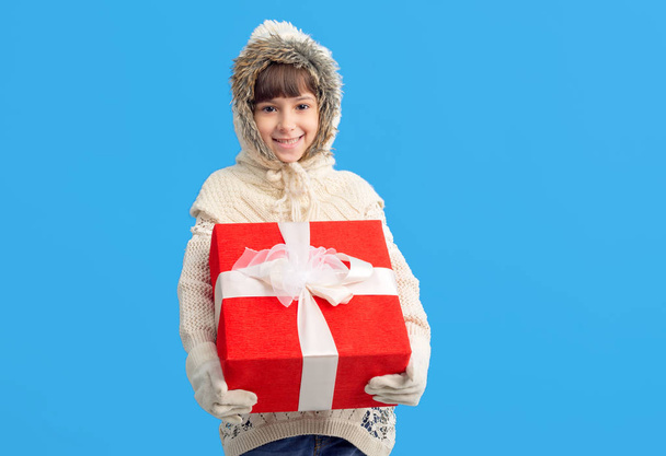 Kislány téli ruhákat, egy piros ajándék doboz a kezek-ra egy kék háttér. Karácsonyi ajándékokat. - Fotó, kép