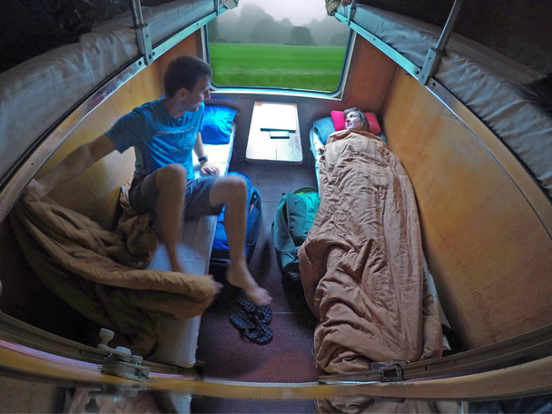 Mężczyzna podróżnik wstaje z łóżka, a jego dziewczyna odpoczywa podczas jazdy pociągiem po malowniczej okolicy w Wietnamie. Para młodych turystów biorąc podkładów pociąg do podróży w Azji. - Zdjęcie, obraz