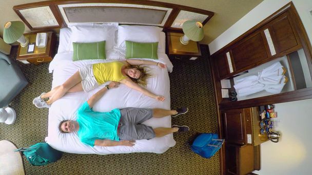 Πάνω προς τα κάτω: Παιχνιδιάρικο νεαρό ζευγάρι για χαλαρωτικές διακοπές πτώση επάνω το άνετα, king size κρεβάτι σε hostel τους. Καυκάσιος νεόνυμφους μεταφέρουν τσάντες ταξιδίου τους στο διαμέρισμά τους και να πηδούν πάνω στο κρεβάτι. - Φωτογραφία, εικόνα