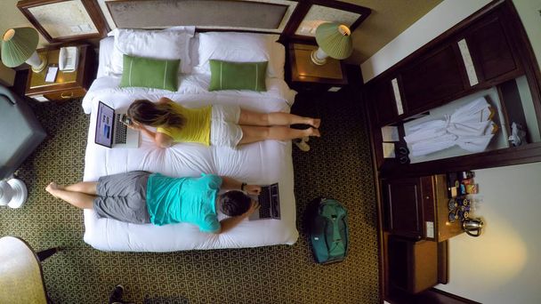 トップダウン: 若い彼らのホテルの部屋の大きなベッドの上に横たわっている間、コンピューターでの作業をカップルします。ベトナムの休日の男性と女性のデジタル遊牧民を入力し、楽しい中に自分のラップトップに取り組んで. - 写真・画像