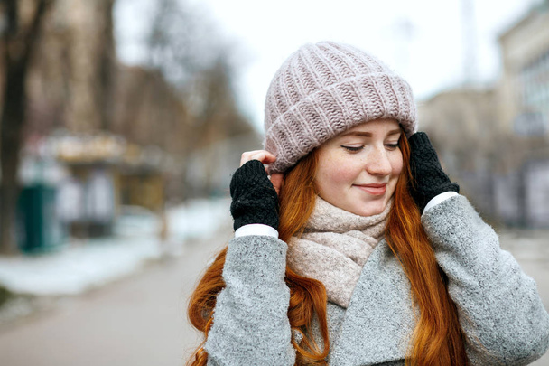 Closeup shot of smiling ginger model with long hair wearing knitted cap and scarf walking at the city. Space for text - Fotó, kép