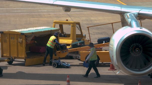 Ground staff collecting passengers' luggage coming out of the international airplane's fuselage. Airport employees in reflective vests moving travel bags from conveyor belt to yellow luggage truck. - Photo, Image
