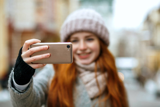 Joyful red head woman in winter apparel taking selfie on her mobile phone. Space for text - Фото, изображение