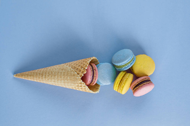 Waffle cone with colorful macaron or macaroon on blue pastel background top view. Flat lay composition - Foto, Bild