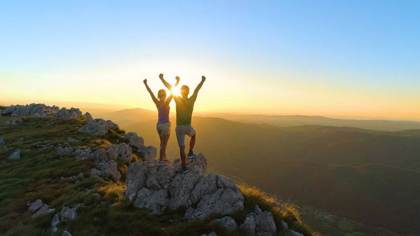 DRONE, LENS FLARE: Flying over sporty couple outstretching arms after a successful hike in the scenic mountains in Slovenia. Cheerful young tourists outstretching arms on a golden summer evening. - Photo, Image