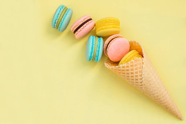 Waffle cone with colorful macaron or macaroon on yellow pastel background top view. Flat lay composition - Foto, immagini