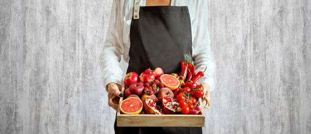 girl holds wooden tray with fresh red vegetables and fruits on grey background. Healthy eating vegetarian concept. - Photo, image