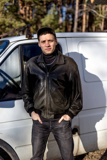 Молодой парень с криминальной внешностью в черной кожаной куртке стоит рядом со старым белым фургоном
. - Фото, изображение