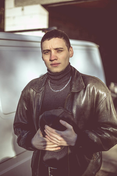 A young guy of criminal appearance in a black leather jacket stands near an old white van. - Φωτογραφία, εικόνα