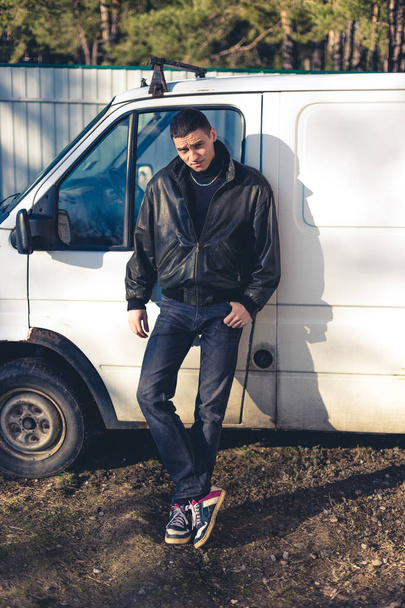 Un giovane ragazzo dall'aspetto criminale con una giacca di pelle nera si trova vicino a un vecchio furgone bianco
. - Foto, immagini