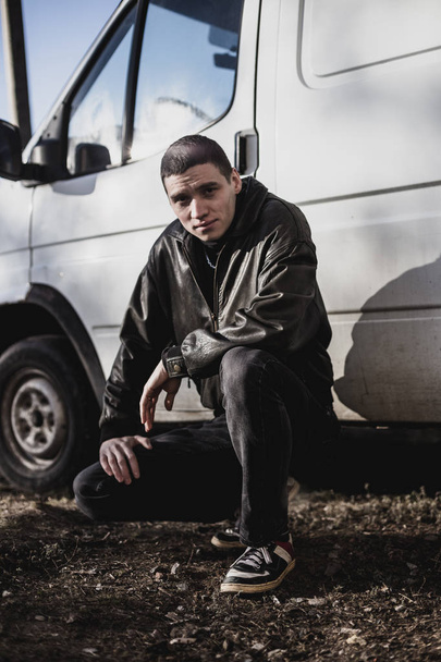 Un jeune homme d'apparence criminelle dans une veste en cuir noir se tient près d'un vieux van blanc
 - Photo, image