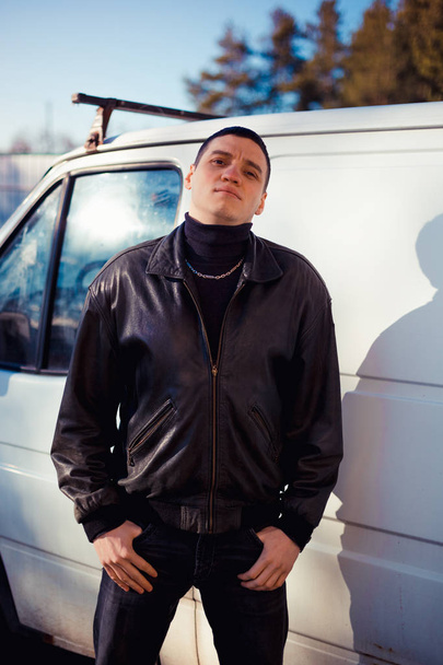 Un giovane ragazzo dall'aspetto criminale con una giacca di pelle nera si trova vicino a un vecchio furgone bianco
 - Foto, immagini