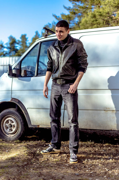 Genç bir adam bir siyah deri ceketli suç görünüm eski bir beyaz van duruyor. - Fotoğraf, Görsel