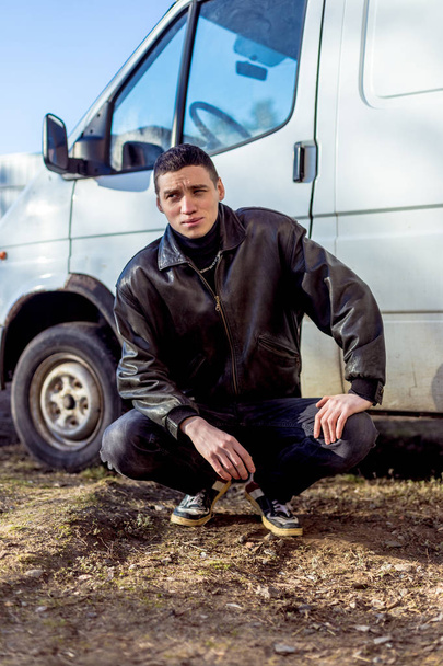 Een jonge kerel van criminele verschijning in een zwart lederen jas staat in de buurt van een oude witte bestelwagen. - Foto, afbeelding